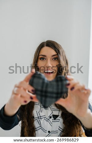 Picture of a small gray heart in hands, female holds handmade sewn soft toy, woman with Valentine gift against the window, happy girl smiling, conceptual image of health care or love.