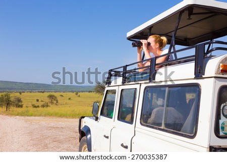 Young blond lady on safari standing in open roof jeep observing wild animals through binoculars. Royalty-Free Stock Photo #270035387
