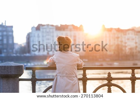 A young woman is looking at the sunset over a river in the city Prague with the old buildings in the background. Soft spring backlit. Color toned image. Royalty-Free Stock Photo #270113804