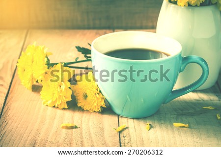 soft focus photo,blue cup of coffee on wooden floor with yellow flower in white pot and good morning paper note on morning sunlight. vintage color tone. 