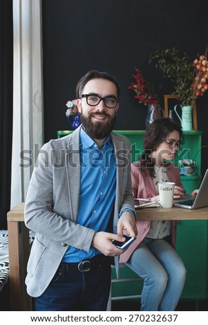 Young businessman in glasses with smartphone over woman working on laptop on background