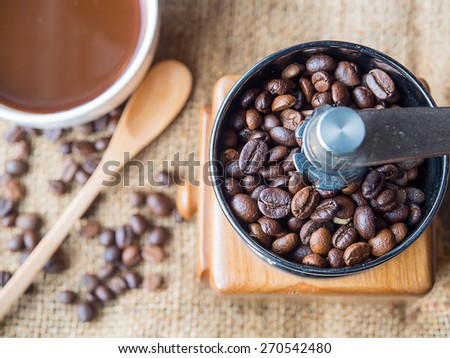 A photo of the roasted coffee beans in a coffee grinder with blur coffee beans , fresh coffee , wooden spoon on sack background.