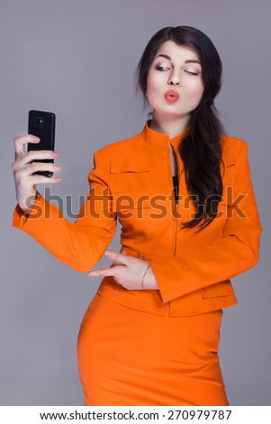 Beautiful brunette caucasian woman in orange stylish business suit make selfie on the phone. grey neutral background