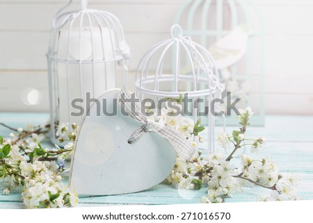 Background with white spring flowering branches of trees, decorative heart  and candles in  bird cages in ray of light on turquoise painted wooden planks. Selective focus is on heart. Place for text. 