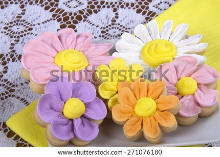 Fancy home made spring flowers sugar cookies with royal icing petals piped on. Served on a square plate with yellow napkin underneath. 