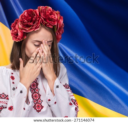 Patriotic concept. Sorrowful beautiful young girl in Ukrainian national suit covers her face with her hands against Ukrainian flag background