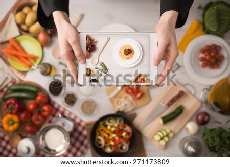 Cook's hands holding a touch screen tablet close up, kitchen table with food ingredients, vegetables and utensils on background, top view Royalty-Free Stock Photo #271173809