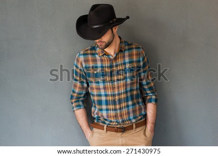 Cowboy couture. Portrait of young man wearing cowboy hat and looking down while standing against grey background 