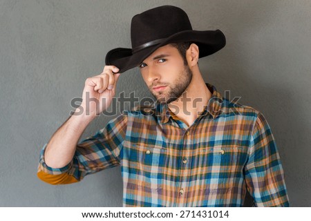 Cowboy style. Handsome young man adjusting his cowboy hat and looking at camera while standing against grey background  Royalty-Free Stock Photo #271431014