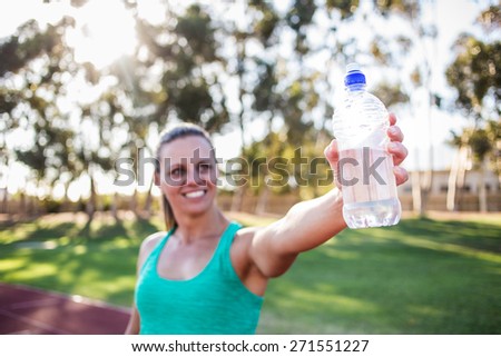 Close up wide angle view of a female fitness athlete holding up a bottle of water with a smile on her face. Back light natural light.