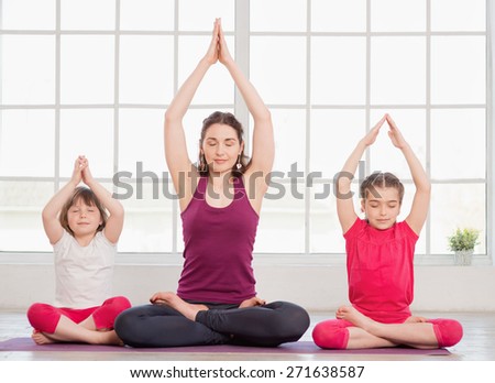 Young mother and daughters sitting in lotus position and doing yoga exercise in fitness studio with big windows on background