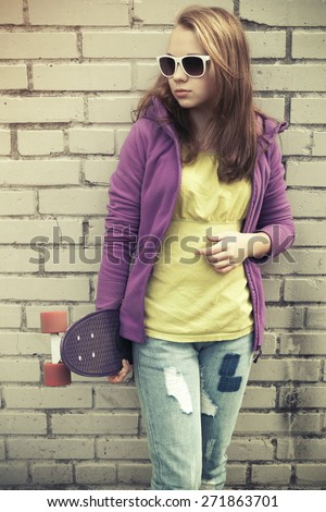 Blond teenage girl in jeans and sunglasses holds skateboard near gray urban brick wall, vintage cold green tonal correction, instagram style photo filter effect