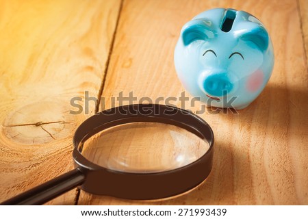 soft focus photo of blue piggy bank on wooden floor with magnifying glass on morning sunlight. vintage color tone.