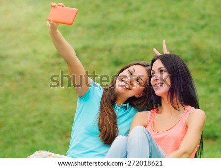 two happy women friends laughing and sharing social media pictures in a smart phone on picnic at the park, lifestyle concept