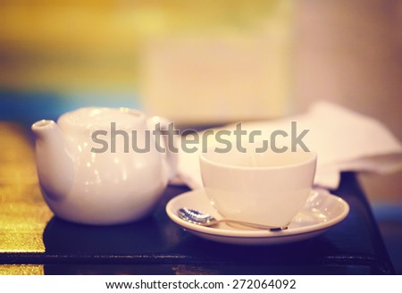 White teapot and cup on a plate with spoon on table near the window in restaurant cafe with evening night lights, Instagram filters used with vintage retro effect, soft focus, blurry background
