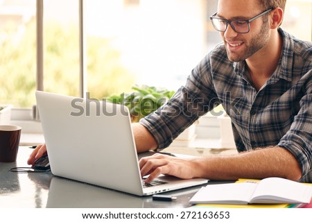 Happy young man, wearing glasses and smiling, as he works on his laptop to get all his business done early in the morning with his cup of coffee Royalty-Free Stock Photo #272163653