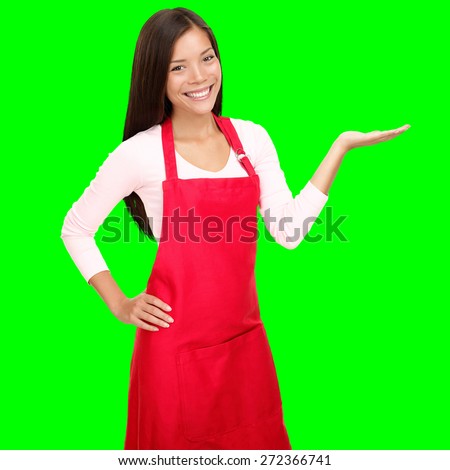 Small shop owner showing empty copy space in red apron. Woman smiling happy presenting with open hand palm. Friendly multiracial Asian Caucasian female model isolated cutout on green background.