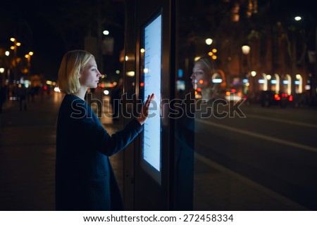 Smart city technology concept, young woman using modern urban application of bus stop in Barcelona for find out schedule of transport, female touching big digital screen which reflecting light Royalty-Free Stock Photo #272458334