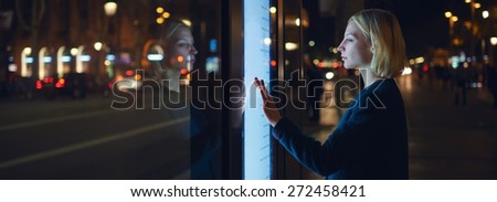 Panoramic picture,female using automated teller machine with big digital screen while standing in night city out-of-focus lights,woman verifies account balance on banking application via modern device