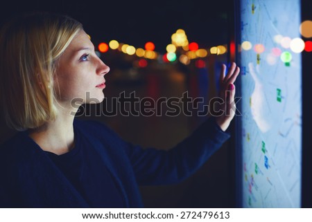 Young female tourist using smart city gadget to get direction in Barcelona central, female in night city standing front big digital screen with city map routes and locations shown on it