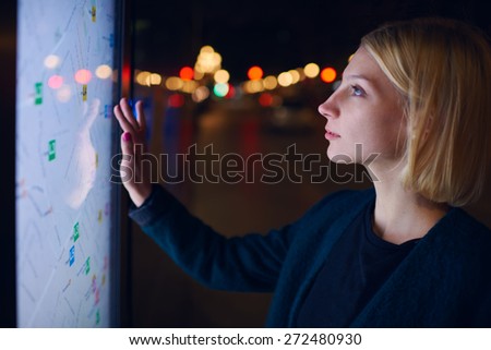 Gorgeous young woman standing front big digital screen with basic city street map data, female touching sensitive display of modern smart city bus stop for check her location into the night