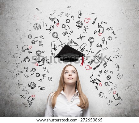 Young beautiful business woman is thinking about education at business school. Drawn business icons over the concrete wall. Graduation hat. Royalty-Free Stock Photo #272653865