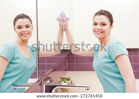 Total reflection. Young woman standing in front of mirror and wiping it with help of white cloth