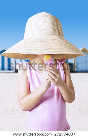 Closeup of little girl wearing a big hat and swimwear on the beach, eating ice cream