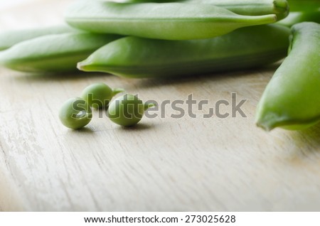 Three young green peas with stalks intact, extracted from a pea pod, with a pile of closed pea pods piled behind them on wooden surface.