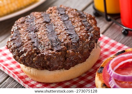 A juicy barbecued hamburger with grill marks on a rustic picnic table with tomato, onion, mustard, ketchup, and corn.