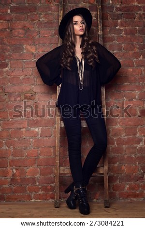 Fashion style portrait of young trendy girl wearing black clothes: blouse, trousers, hat and boots. Model posing and standing near brick wall and wooden ladder.