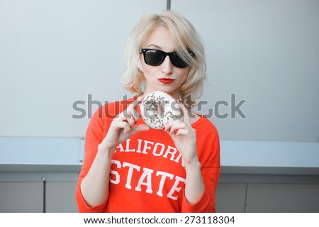 portrait outdoors young beautiful blonde girl wearing black glasses sun with bright makeup, red lips, a smile on his face and a sweet donut and phone in hand, makes Selfie