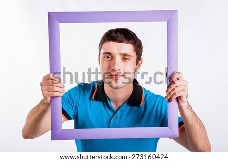 Crazy picture. Handsome young man in shirt holding picture frame in front of his face and smiling while standing against grey background