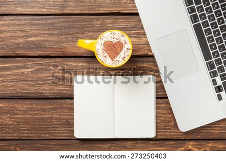 Cup of cappuccino with heart shape and notebook near laptop on wooden table.