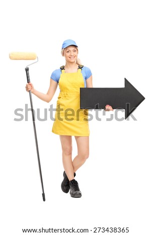 Full length portrait of a female painter in a yellow uniform holding a paint roller and a big black arrow pointing right isolated on white background