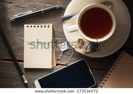 "Good morning" is written on small notepad with a cup of tea, pen, pencil and cellphone on rustic wood background with low key scene Royalty-Free Stock Photo #273536972