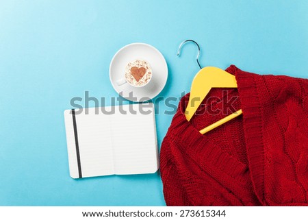 Cup of cappuccino with heart shape and notebook with hanger on blue background.