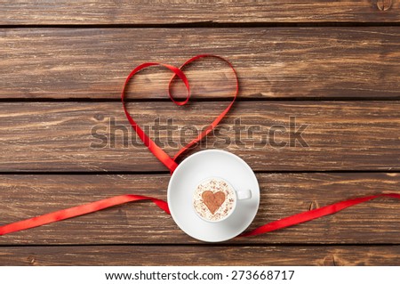 Cup of cappuccino with heart shape and red ribbon on wooden background.