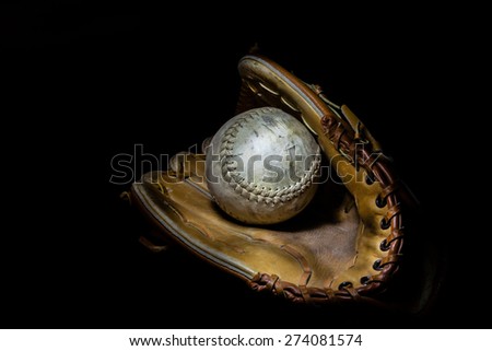 A worn softball sits inside an old baseball glove on a solid black background.  Image was lit by using a lightpainting technique.