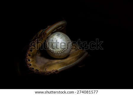 A worn softball sits inside an old baseball glove on a solid black background.  Image was lit by using a lightpainting technique.