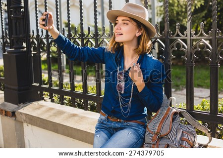 young stylish smiling pretty woman taking pictures selfie her phone, dressed in denim shirt and jeans, with hat and backpack, sunny day, good weather, city street, cool accessorize, vacation europe