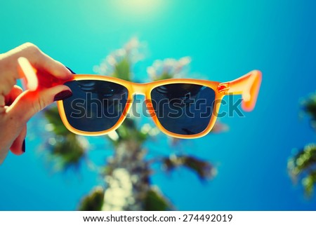 Female hand holding colorful sunglasses against palm tree and blue sunny sky, summer vacation holidays concept, first person shot, looking though glasses, filtered image