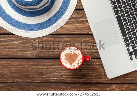 Cup of cappuccino with heart shape and hat near laptop on wooden table.