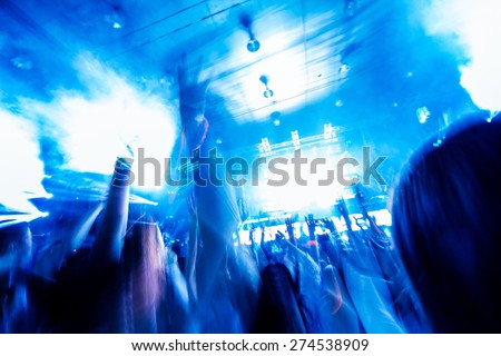 Picture of concert, music festival,party in nightclub, dance floor, disco club, many people standing with raised hands up. Blurred motion in night light rays.