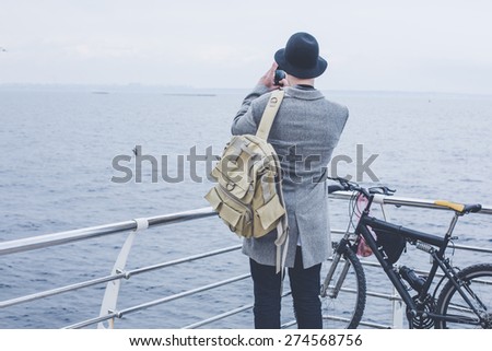 handsome young hipster guy in hat with bike taking picture of the sea with his smart phone wearing backpack on wood floor back view
