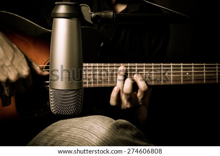 male musician playing acoustic guitar behind condenser microphone in recording studio