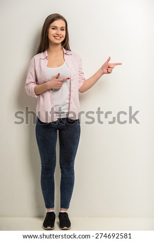 Young smiling beautiful girl in full length showing empty open space for text on grey background.