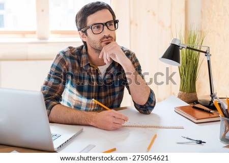 Waiting for inspiration. Thoughtful young man holding hand on chin and looking away while sitting at his working place Royalty-Free Stock Photo #275054621