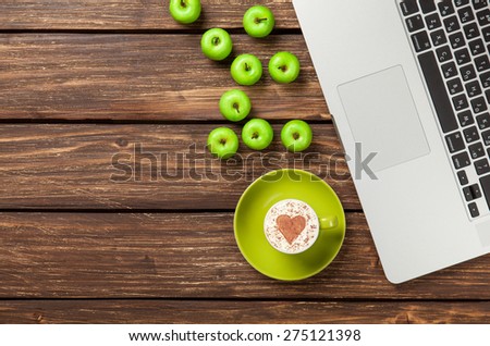Cup of cappuccino with heart shape and apples near laptop on wooden table.