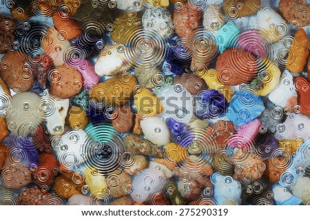 A close up of water covered, multicolored pebbles and stones in a river bed or pond with rain ripples